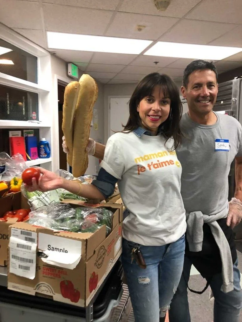 A woman and man smiling in a food pantry, with the woman holding a tomato and the man wearing a gray shirt with a blue name tag. The woman is wearing a white T-shirt that says 'maman je t'aime' and gloves.