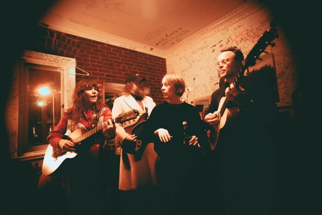 Four people singing and playing guitars in a cozy, dimly lit room with graffiti-covered walls.