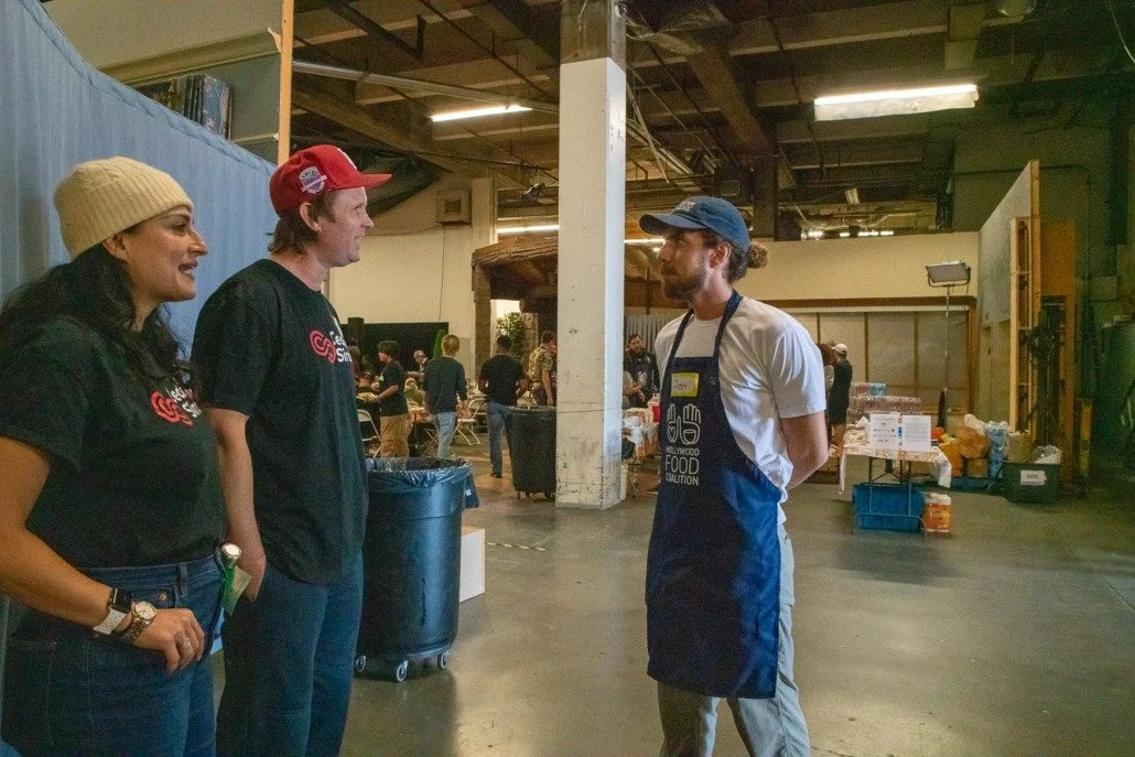 Three people, two women and one man, talking to a man wearing an apron at a community food event in an industrial-style space.
