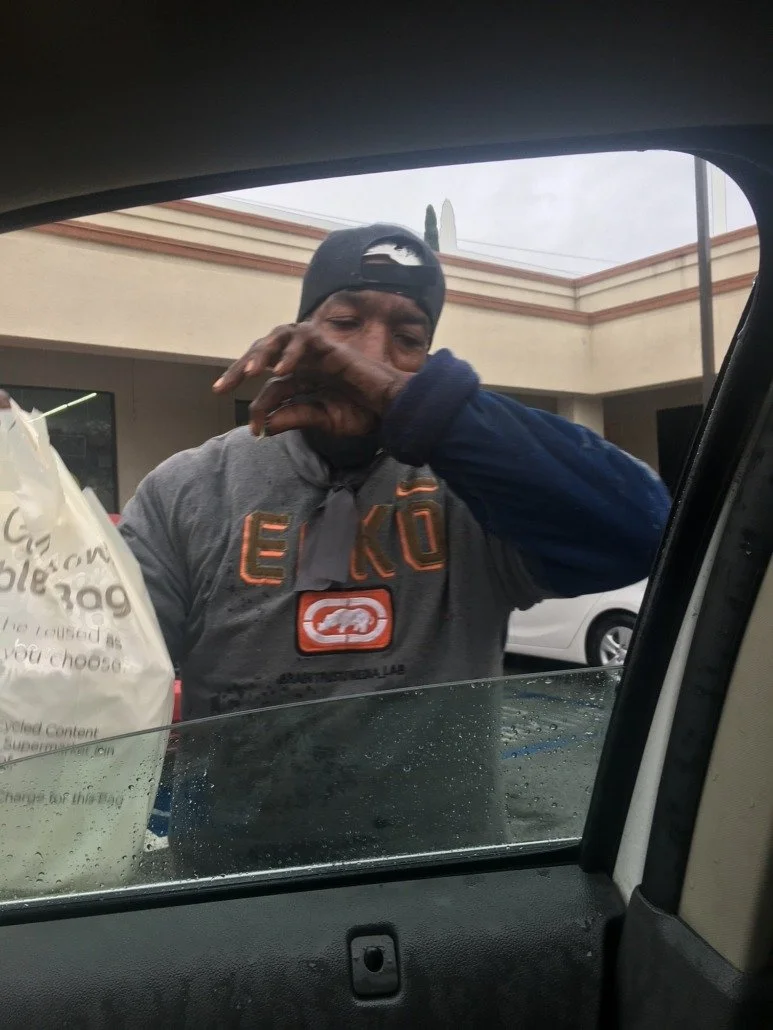 A man wearing a black baseball cap and a black apron is reaching out to the camera through a car window, with a beige building and cloudy sky in the background.