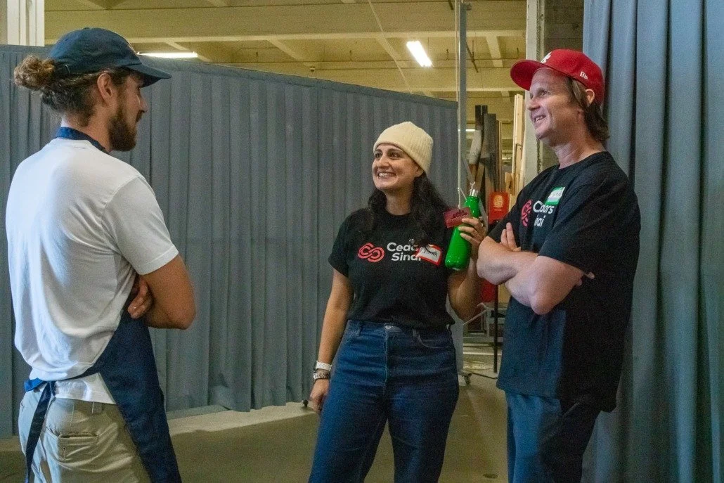 Three people standing and talking in a work area, two of them wearing black Cooks Aid shirts, one woman with a white beanie holding a green spray bottle, and two men with caps, one with a red cap and the other with a dark cap, all smiling.