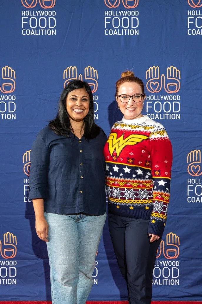 Two women standing in front of a blue backdrop with the Hollywood Food Coalition logo, smiling for the camera.