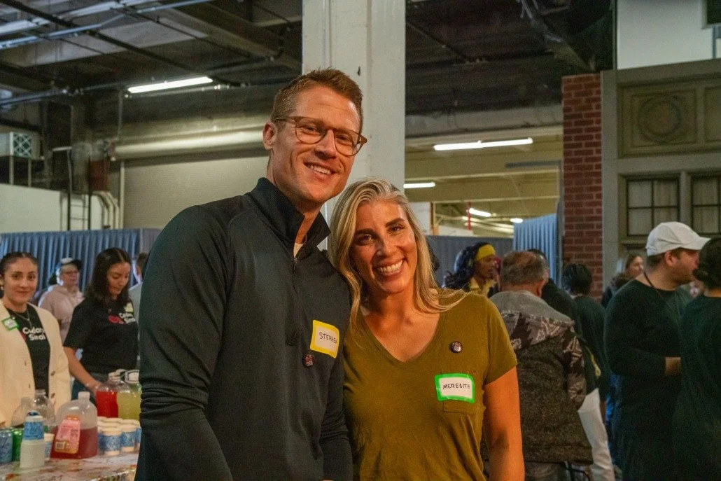 A man and woman smiling at the camera at an indoor event. The man is wearing glasses and a black jacket, and the woman has blonde hair and is wearing a green shirt. They have name tags that read 'Stpace' and 'Meredith'. There are other people and boo