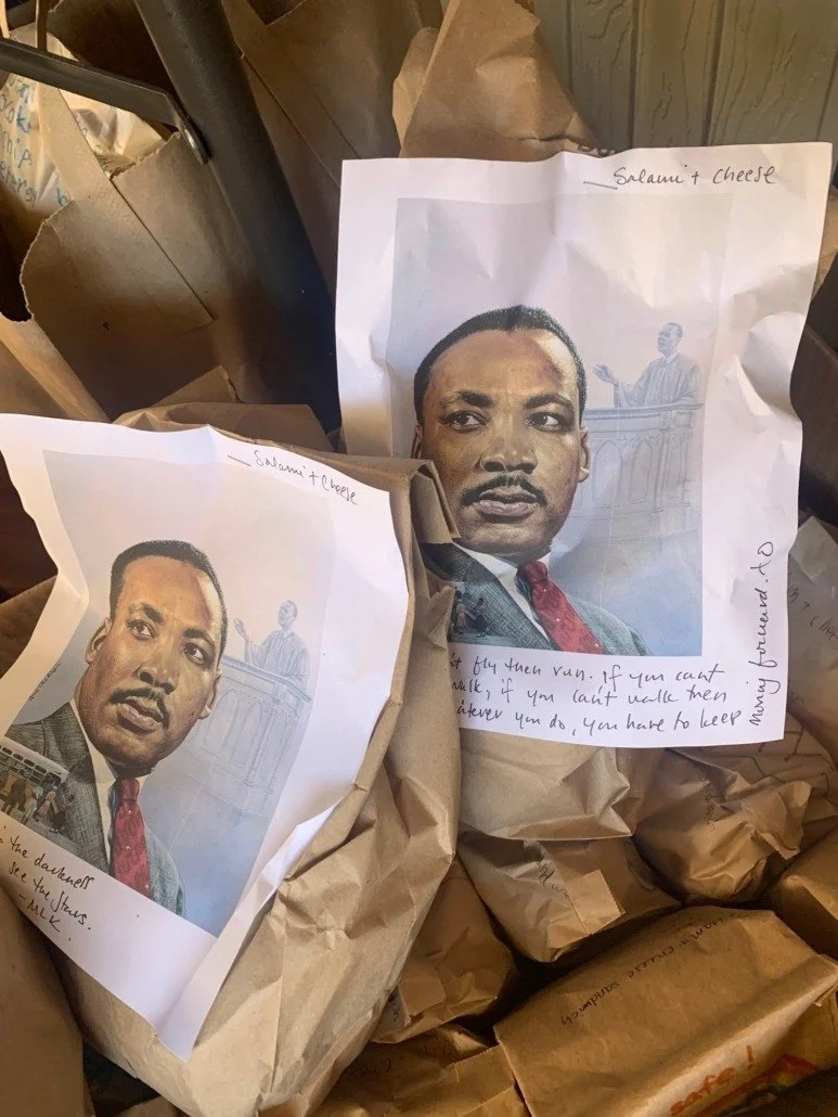 Two paper bags with printed photos of Martin Luther King Jr. attached to them, surrounded by brown paper packaging material. The photos include handwritten notes referencing a quote and the words 'Salami + Cheese'.