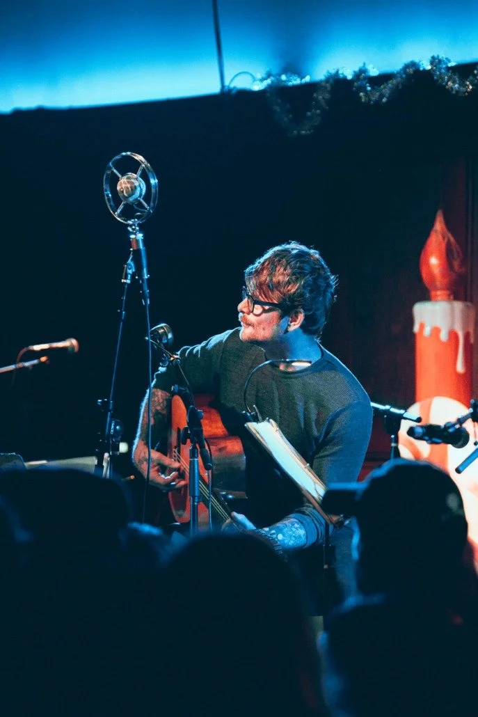 A musician performs on stage playing an acoustic guitar, wearing glasses and a long-sleeved shirt, with a music stand in front and a microphone nearby, in a dimly lit venue with an audience.