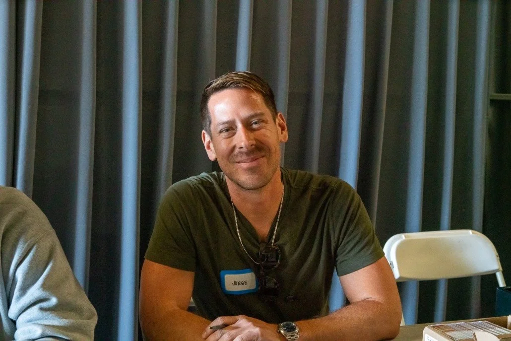 A man with short brown hair and a beard smiling, sitting at a table with a gray curtain background, wearing a green t-shirt, a watch, and a name tag that says 'Jorge'.