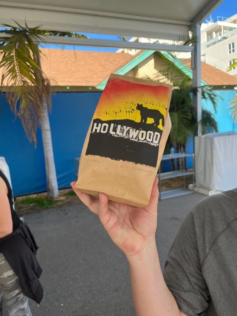 A hand holding a paper bag with the Hollywood sign styled logo, featuring a silhouette of a dog, against a background of palm trees, a blue fence, and a tent at an outdoor setting.