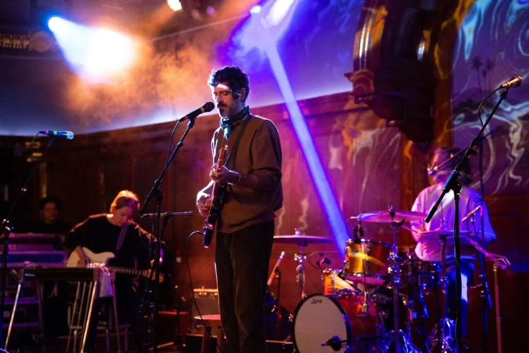 Musicians performing on stage with colorful lighting, including a guitarist at center, a drummer, and keyboardist in the background.