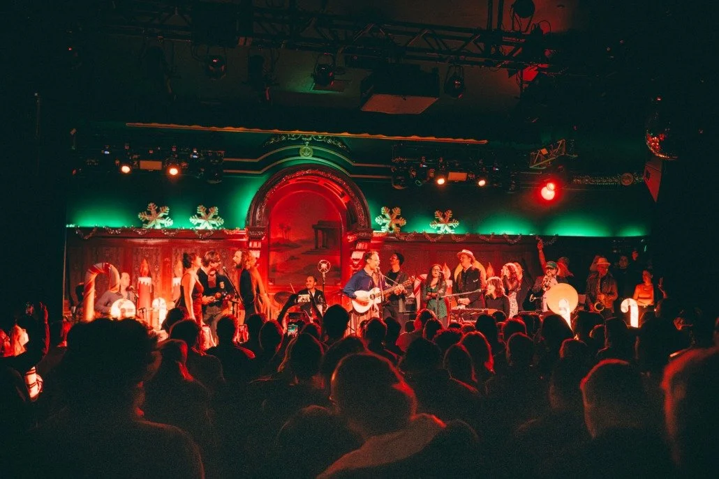 A live holiday concert on stage with musicians playing and singing, decorated with Christmas ornaments and lights, audience watching.