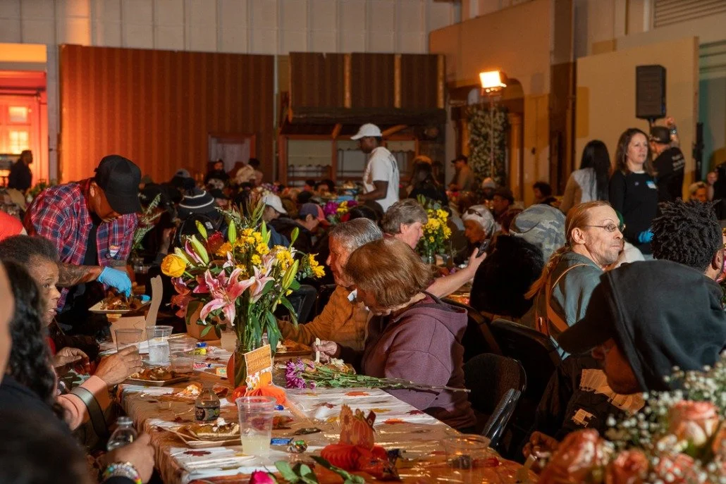 People gathered around a long table decorated with flowers, sharing a meal at an indoor event, with waitstaff serving food in the background.