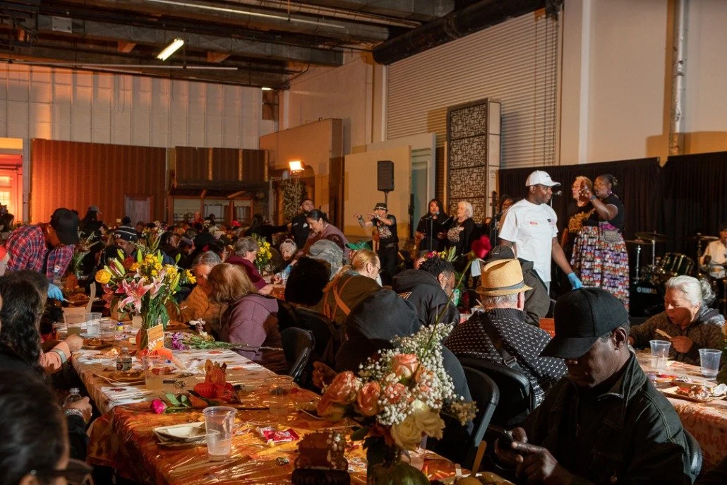 A large indoor gathering with many people seated at tables covered in orange tablecloths, decorated with flowers, and eating. A stage at the far end of the room features performers and speakers, including a woman in a colorful dress speaking into a m