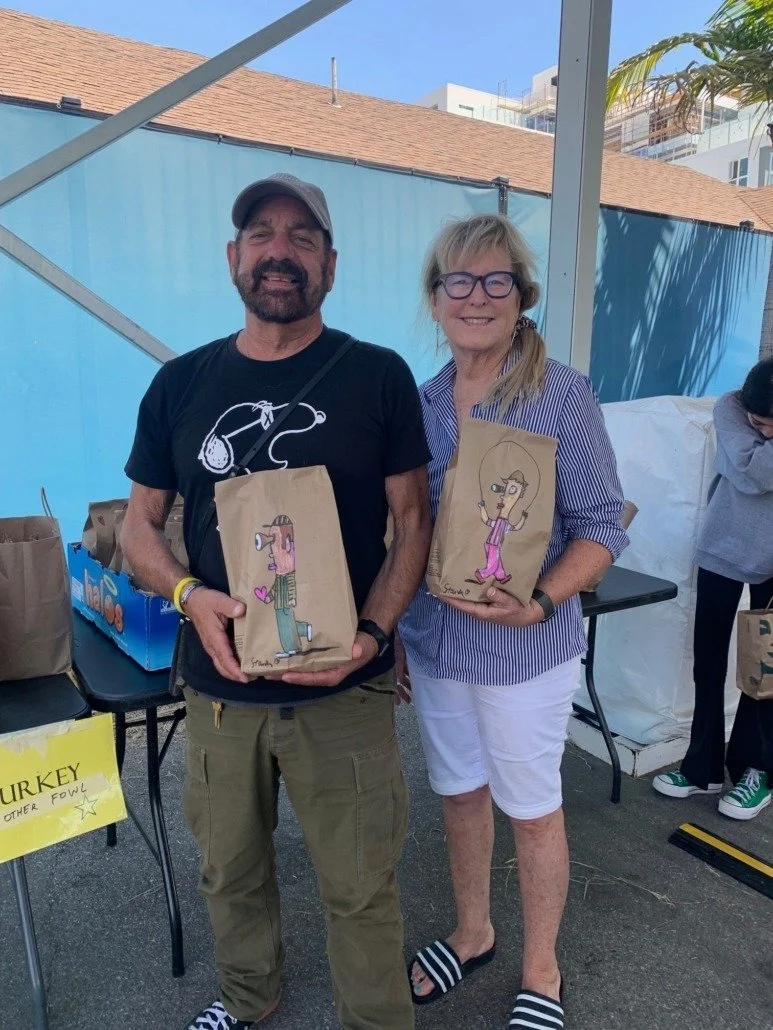Two people standing outdoors, each holding a paper bag with a cartoon drawing. The man on the left is wearing a black t-shirt and cap, while the woman on the right is wearing glasses, a striped shirt, and white shorts. They are smiling and posing for