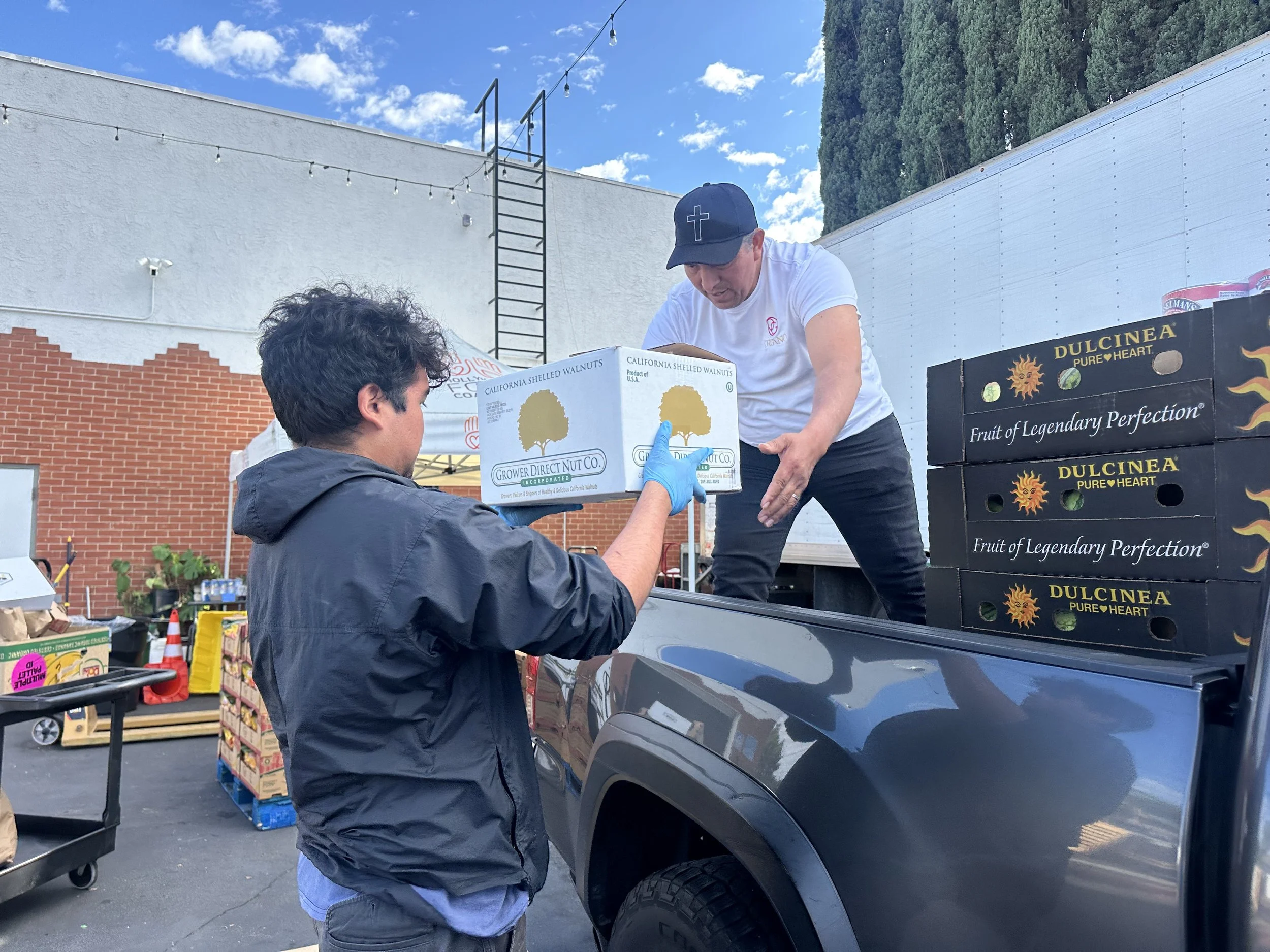 Two men loading produce, including boxes of walnuts and crates of fruit, into a truck at an outdoor market or distribution event.