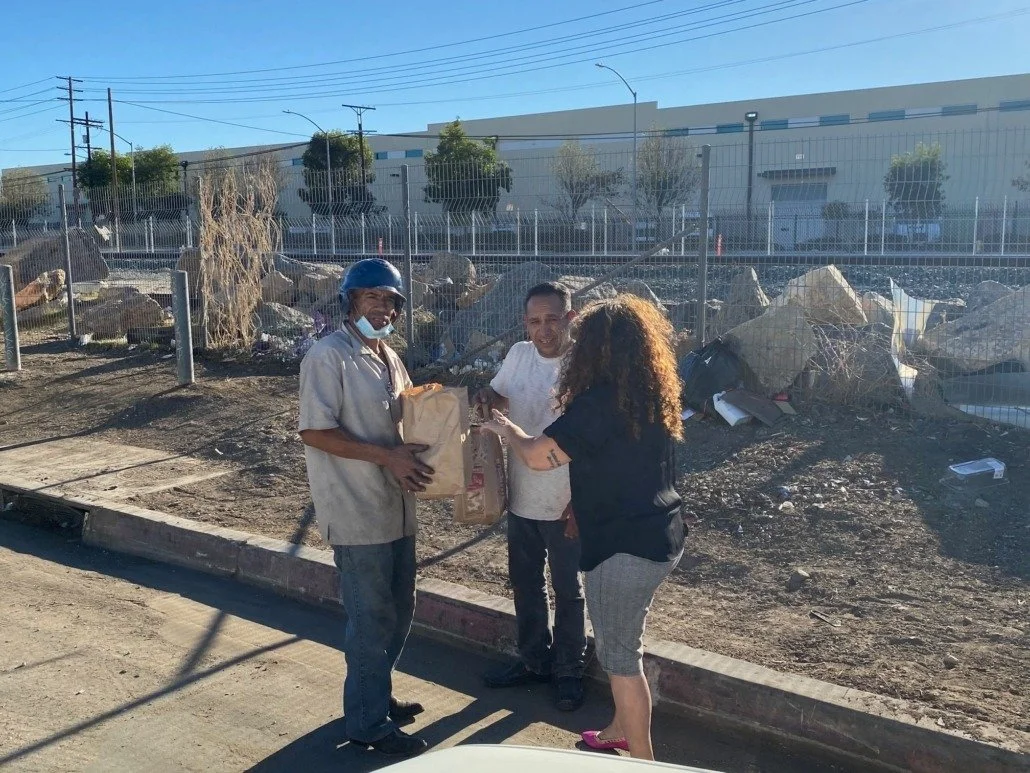 Three people are standing on a sidewalk near a construction or demolition site, with a chain-link fence and rocks behind them. One person is handing over a paper bag to another, and the third person is holding another paper bag. It appears to be a co