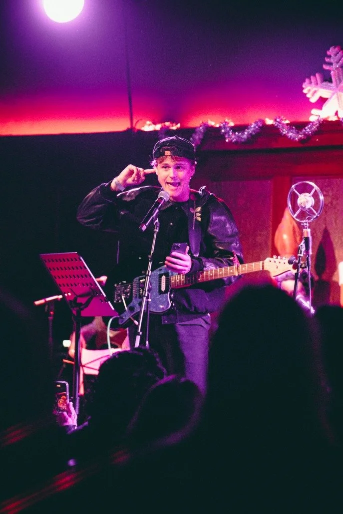 A young male musician performing on stage with a guitar, wearing a black jacket and headband, holding a phone in one hand and speaking into a microphone. Stage decorated with Christmas ornaments and decorations, audience visible in the foreground.