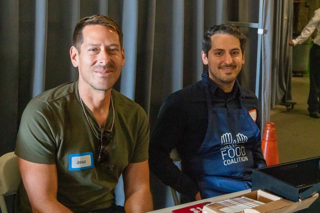 Two men sitting at a table, one wearing a green t-shirt with a name tag that says 'Jorge' and sunglasses hanging from his shirt, the other wearing a black shirt and an apron with the logo 'Holl Food Coalition', indoors with black curtains behind them