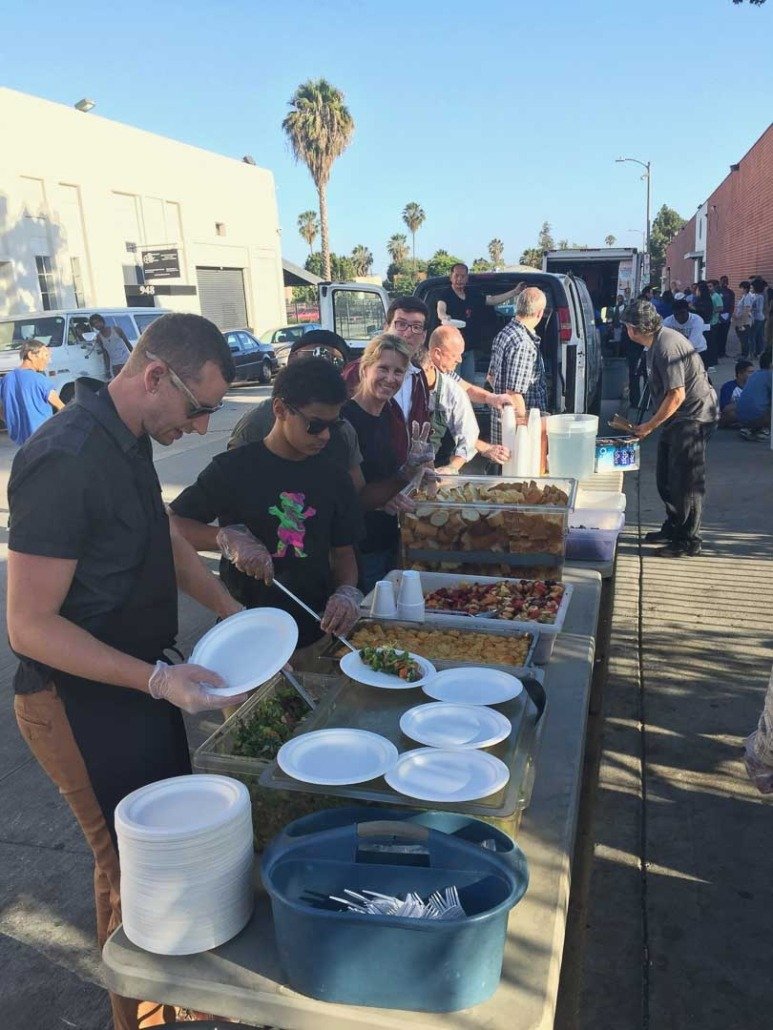 People lining up at an outdoor food distribution or community event, serving themselves food from trays of various dishes on a long table.