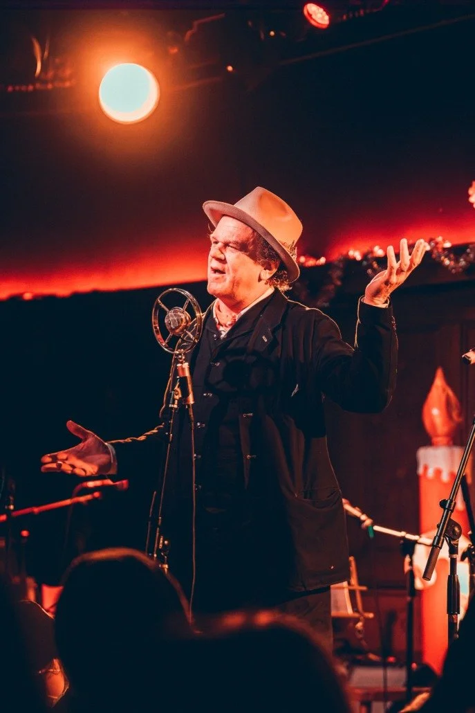 A man performing on stage with a vintage microphone, wearing a tan fedora, black coat, and gesturing with his arms, under warm stage lighting.