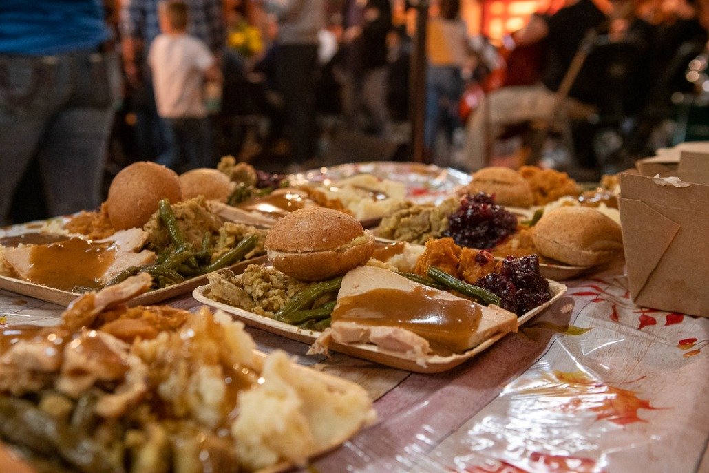 Variety of plates with food, including pies, green beans, mashed potatoes with gravy, and desserts, on a table at a lively outdoor event at night.