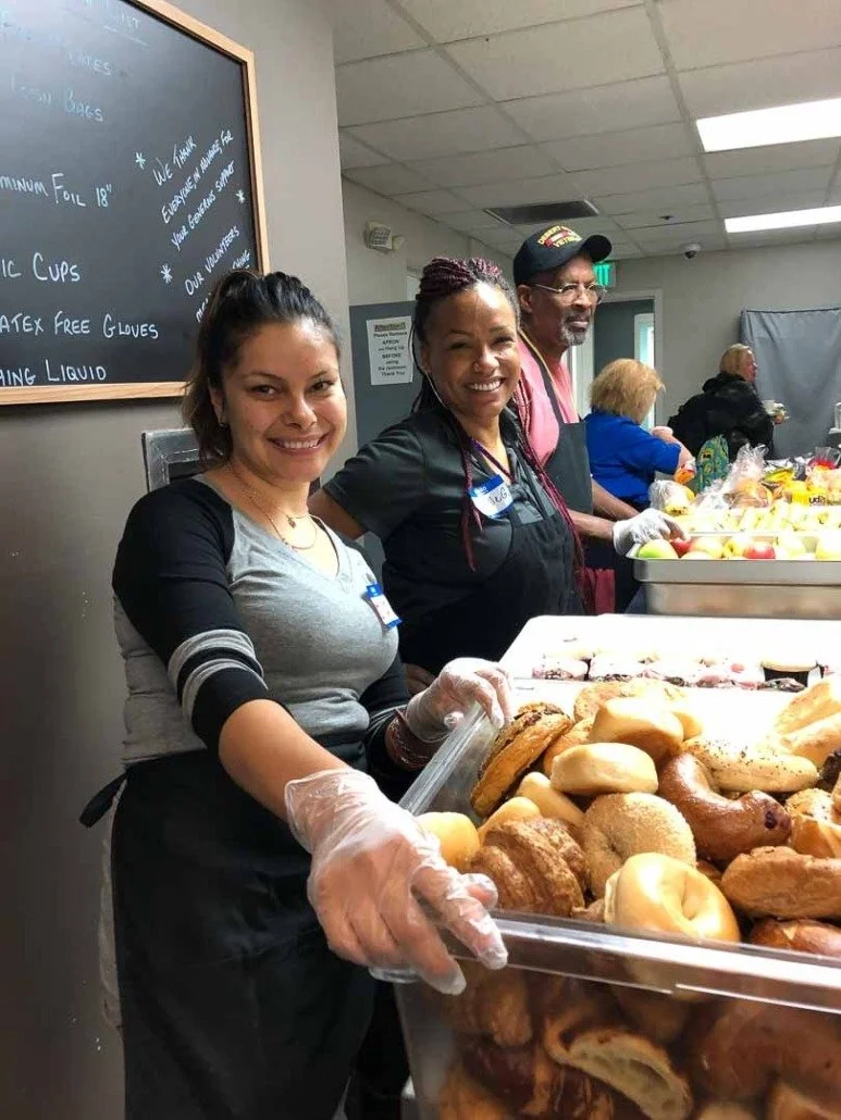 Three people smiling and working at a food serving station with assorted baked goods and fresh produce, including bagels, donuts, and apples, in a community kitchen or cafeteria setting.