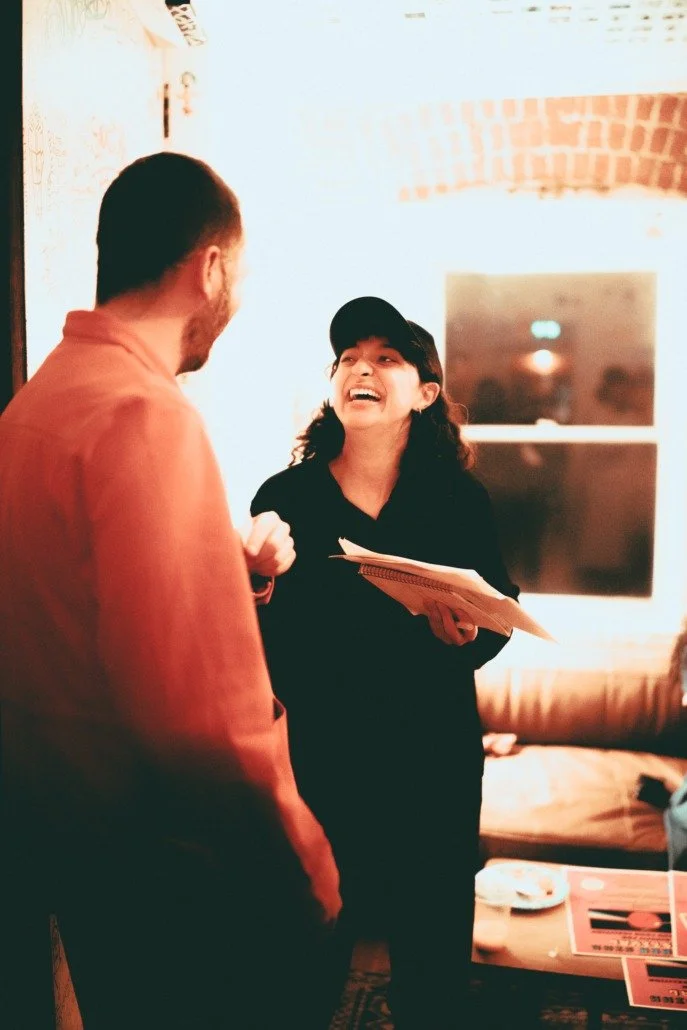 A woman in a black cap and black shirt, holding a notebook, laughing and talking to a man in an orange jacket in a cozy room with a window and a brown couch.