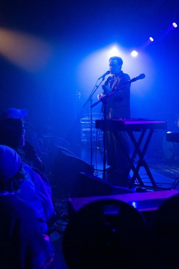A musician performing on stage with a microphone, guitar, and keyboard, illuminated by blue stage lighting, with an audience watching.