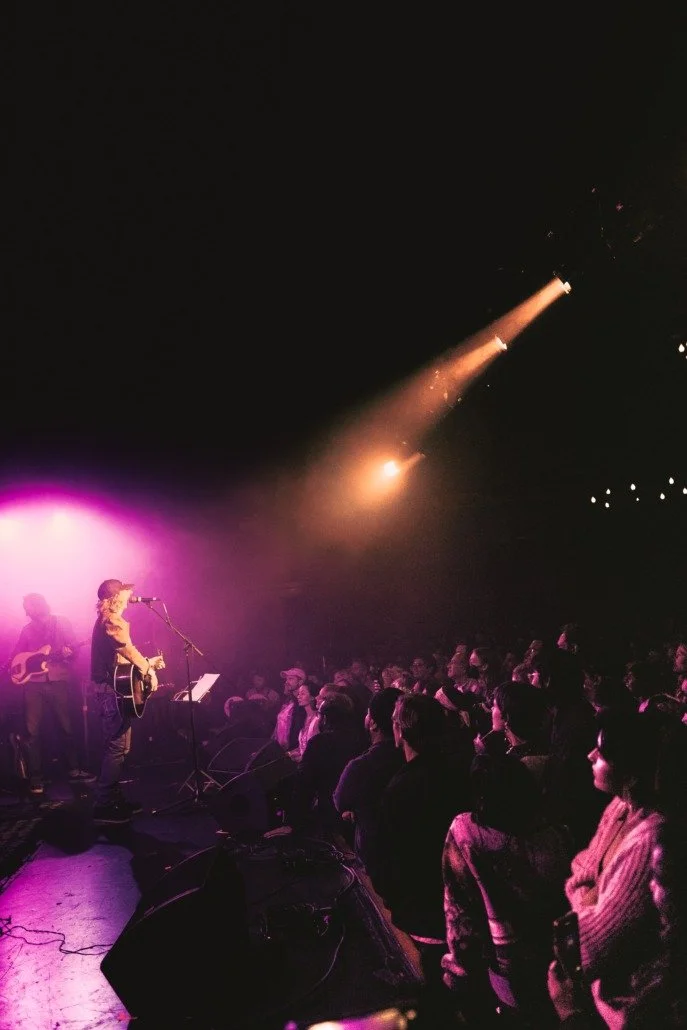 A live music performance with a band and audience in a dimly lit venue, featuring pink and purple stage lighting and a rocket launch visible in the dark sky.