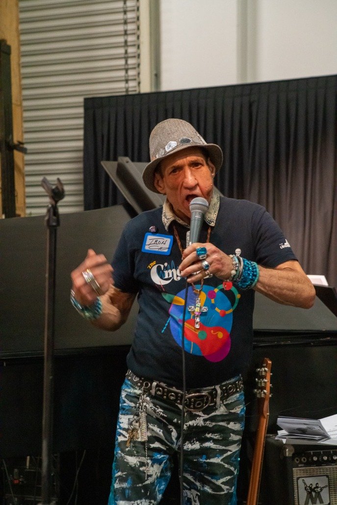 A man wearing a gray hat and black T-shirt with colorful graphics, holding a microphone, and standing in front of a black piano with a dark curtain background.