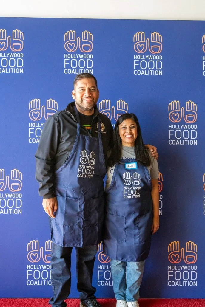 Two people standing in front of a blue backdrop with the Hollywood Food Coalition logo, wearing blue aprons with the same logo, smiling at the camera.