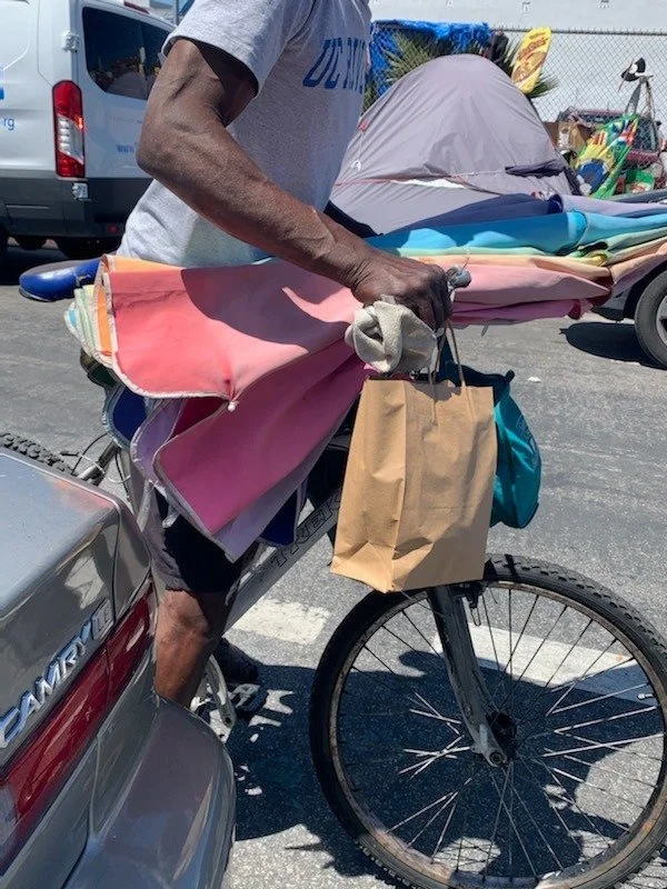 Person on a bicycle loaded with colorful fabric and a paper bag, parked in a lot with vehicles around.