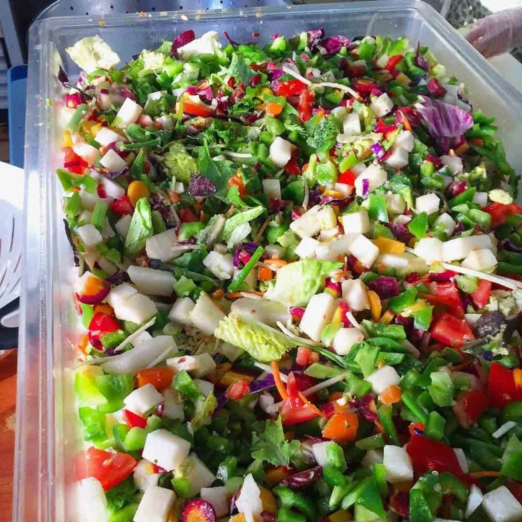 Close-up of a large container filled with chopped mixed salad vegetables including lettuce, tomatoes, cucumbers, bell peppers, radishes, and carrots.