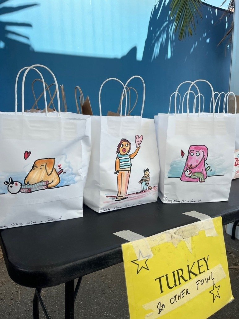 White paper gift bags with cartoon drawings of animals and people, placed on a black table. A yellow sign with black text reading 'TURKEY & OTHER FOWL' is taped to the front of the table.