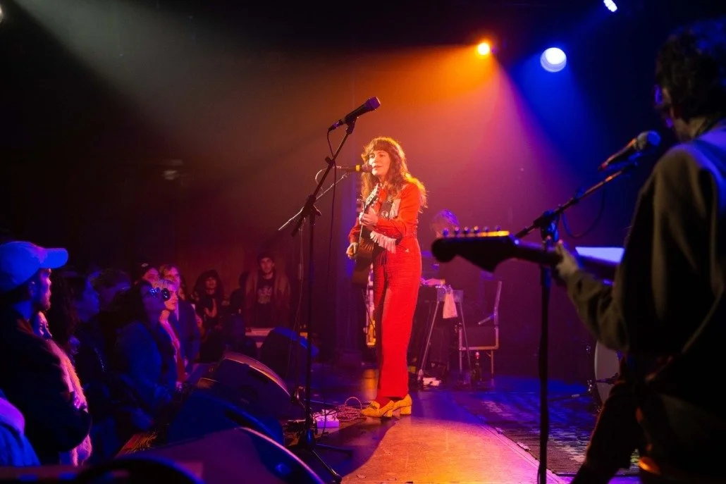 A female singer with curly hair wearing a red outfit is performing on stage with an acoustic guitar. The stage is lit with colorful lights, and an audience is watching her play and sing.