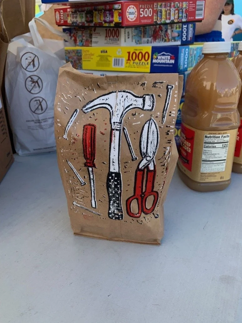 Brown paper bag with a black, white, and red drawing of various hand tools, including a hammer, screwdriver, pliers, nail, and other small tools.