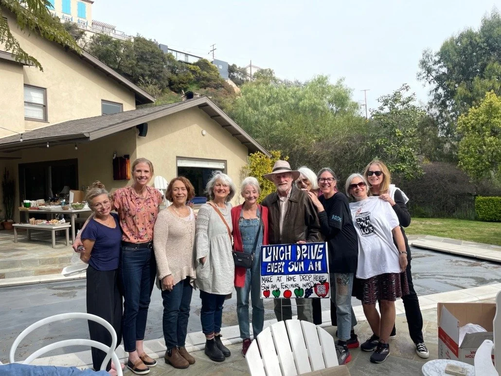 Group of nine women and one man standing outdoors in a backyard, smiling, with a sign that reads "Lunch Drive Every Sun Am" and a table with food in the background.