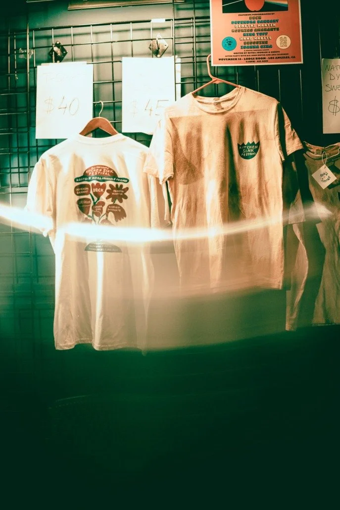 Two T-shirts hanging on a display rack with price signs, one white T-shirt on the left and beige on the right, behind a wire grid wall at a market or stall.