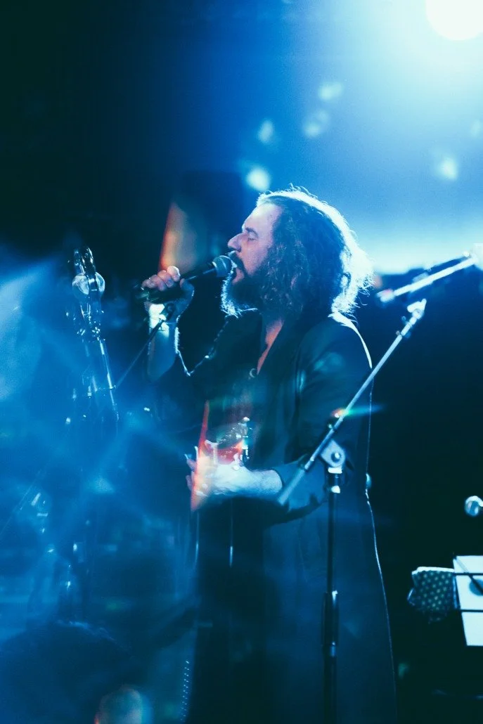 A male singer with curly hair and a beard performing on stage with a microphone, illuminated by blue stage lights.