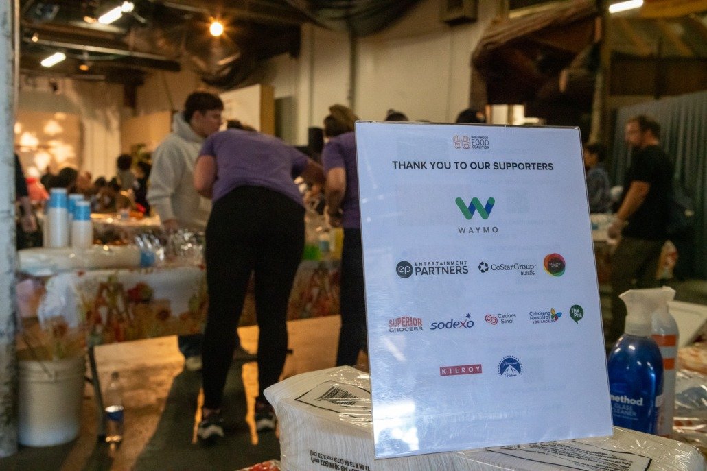A sign on a table thanking supporters of the Waymo food collection event, with logos of various sponsors, in a crowded indoor space with people serving food and multiple tables.
