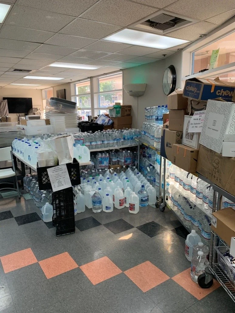 Display of bottled water on shelves and floor in a store or office pantry, with boxes and containers nearby.