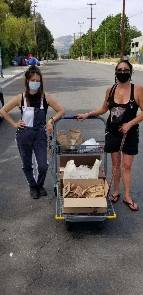 Two women wearing masks standing on a street with a shopping cart filled with paper bags.