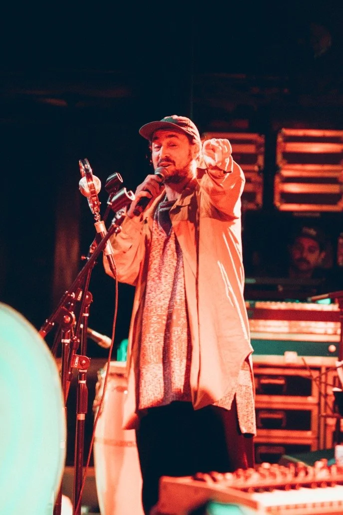 A man wearing a baseball cap and a jacket performs on stage with a microphone, surrounded by musical equipment, in a dimly lit venue.