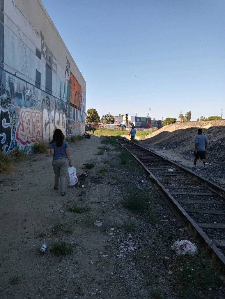 People walking along train tracks beside an urban graffiti-covered wall on a sunny day.