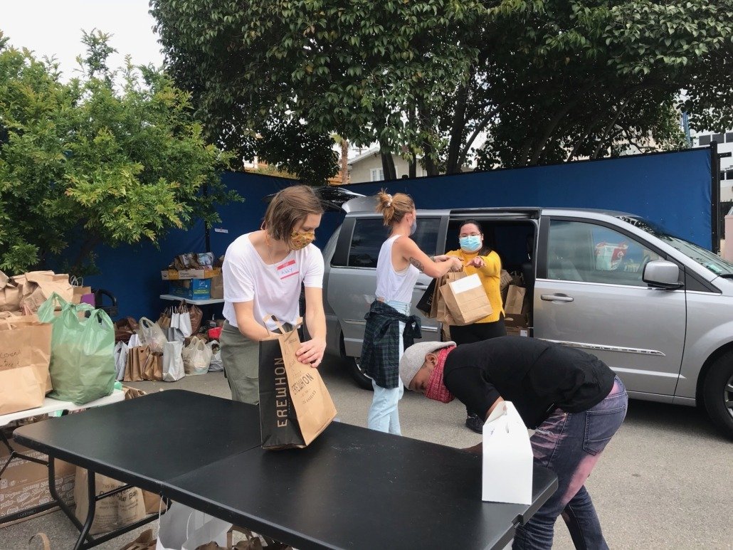 People wearing face masks unloading groceries from a vehicle at an outdoor location with trees and a blue fence.