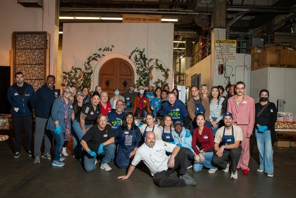 Group of people posing for a photo indoors with a decorative door and plants in the background. Many are wearing blue gloves and some are in aprons, indicating a community or volunteer event.
