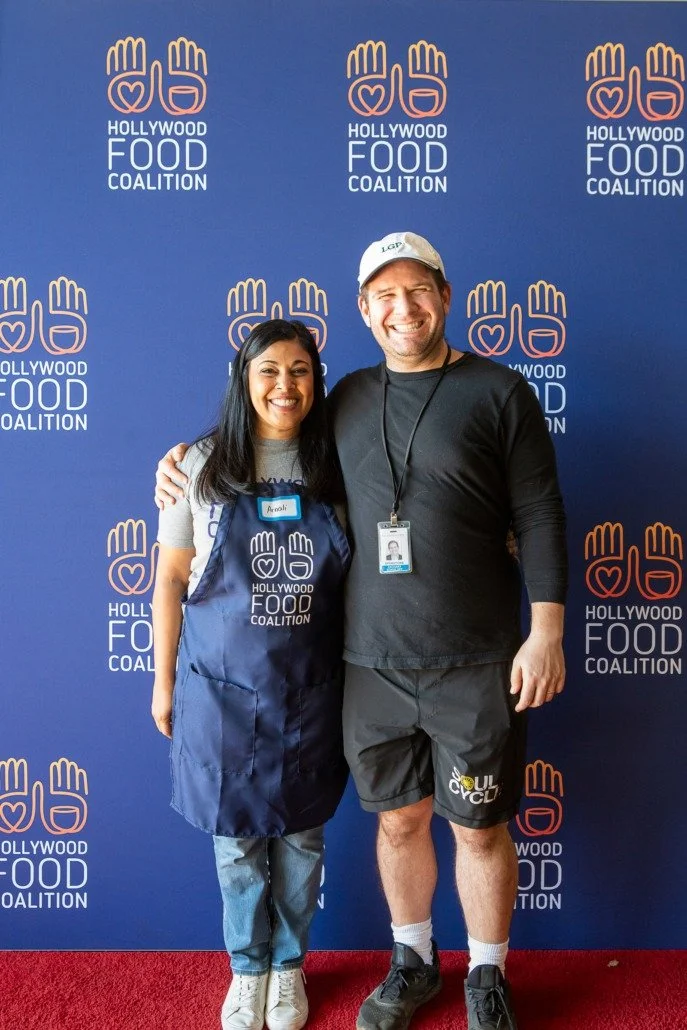 Two smiling people, a woman and a man, standing in front of a blue backdrop with 'Hollywood Food Coalition' logo. The woman wears a gray t-shirt, a blue apron, and has a name tag, while the man wears a black long-sleeve shirt, shorts, and a cap.
