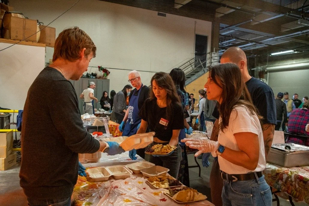 People serving and receiving food at a community meal event in a large indoor space.