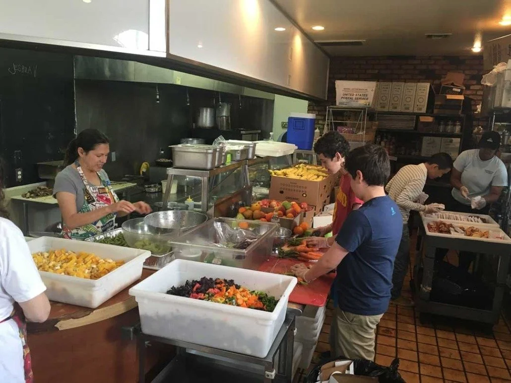 People preparing and packaging food in a kitchen or cafeteria setting with various fresh vegetables and fruits on a countertop.
