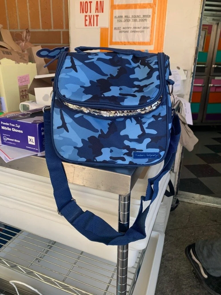 Blue camouflage-patterned school bag on a metal shelf in a store, with a sign that says 'Not an Exit' and some boxes of gloves nearby.