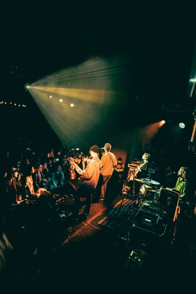 Band performing on stage at a concert with audience in front, stage lights illuminating the scene.