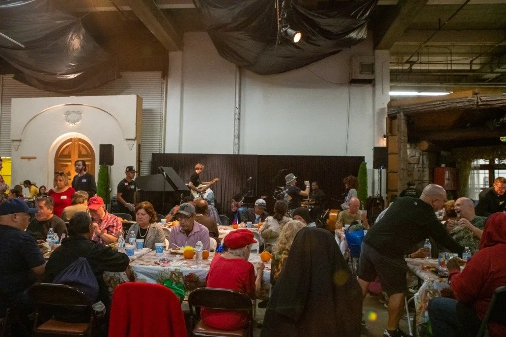 Indoor event with people sitting at tables, a live band performing on stage, some attendees interacting, and festive decorations.